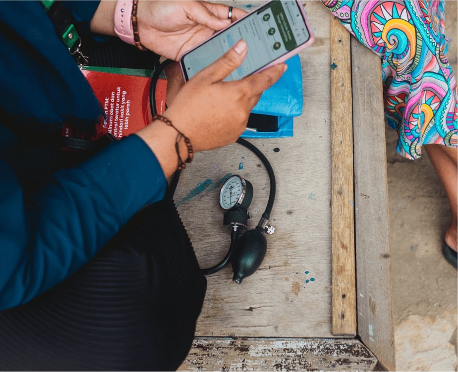 Seorang kader kesehatan sedang melakukan pendataan kesehatan warga setempat melalui aplikasi. Terdapat alat ukur tensi yang digunakan oleh kader untuk mengukur tekanan darah warga dan kemudian dimasukkan ke dalam aplikasi. 