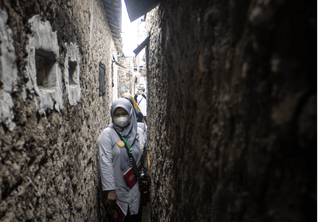 Seorang kader kesehatan yang menggunakan masker sedang menyusuri jalan sempit di antara dua tembok rumah dan sedang dalam perjalanan menjalankan tugasnya. Di belakang nya terdapat dua orang kader kesehatan lainnya yang mengikuti. 